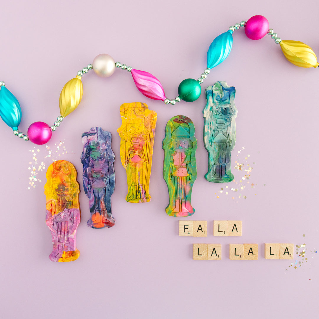 Five nutcracker shaped crayons from Art 2 the Extreme. Each rainbow crayon is shaped like a classic nutcracker figure with vibrant multicolors swirled throughout. The crayon features intricate detailing, including a tall hat, mustache, and uniform-like design, making it a festive and creative holiday gift for children who love coloring. The background is purple with glitter and an ornament garland as photo props.