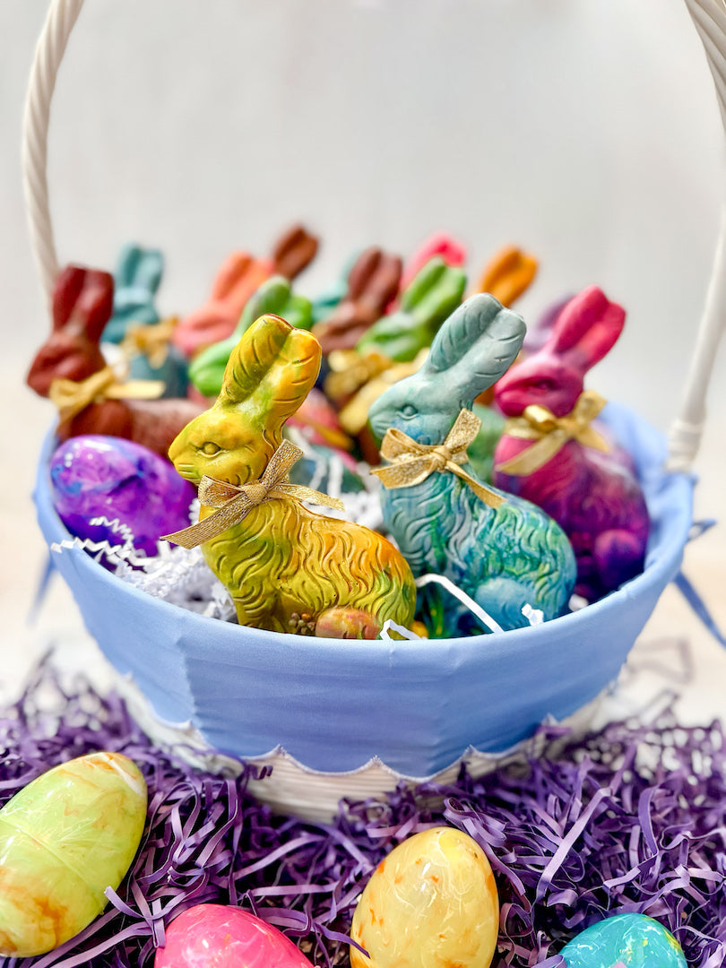 A basket filled with handmade Easter Bunny Original Rainbow Crayons® in various vibrant colors, each tied with a small golden ribbon. The colorful bunny crayons are surrounded by shredded purple paper and marbled Easter eggs, making them a perfect non-chocolate Easter basket filler for kids.