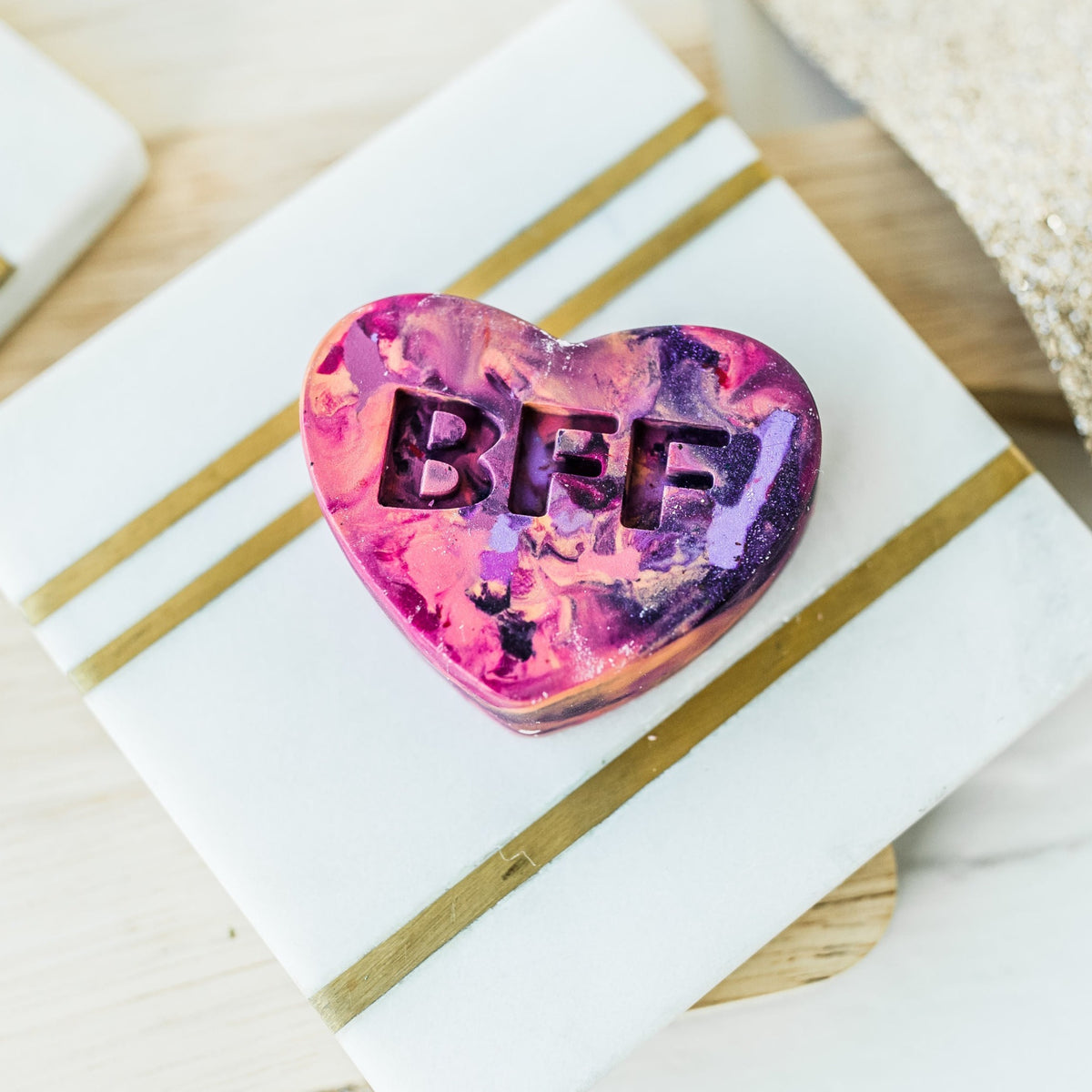 Closeup image of a conversation heart rainbow crayon kids gift from Art 2 the Extreme. Valentine crayon is displayed on a white and gold coaster being used as a photo prop. Valentine crayon favor is a mixture of pink, purple, and peach with the letters BFF engraved on the crayons.