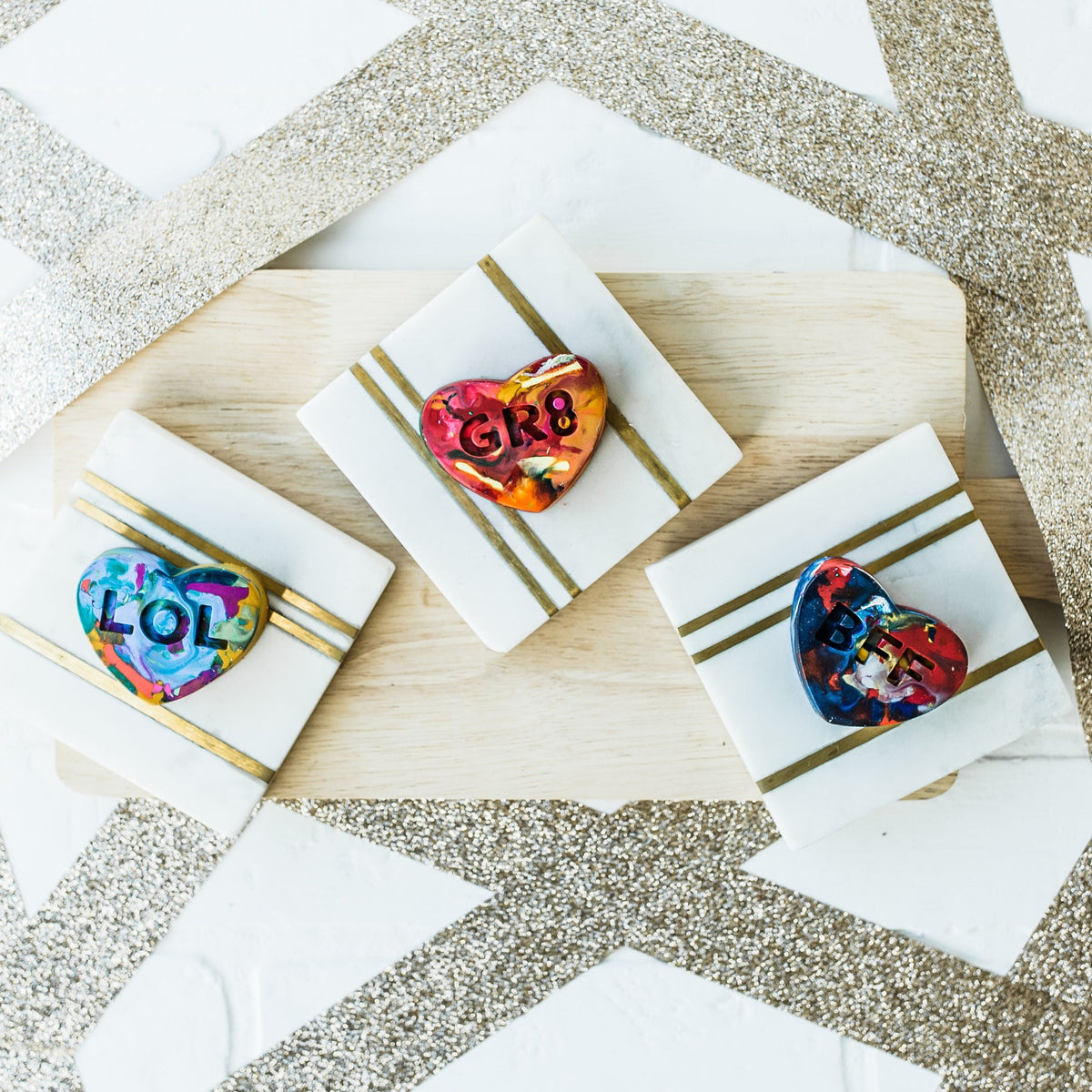 Three multi-colored conversation heart valentine crayons for kids are displayed on a white and gold background with a wooden charcuterie board as a photo prop. 