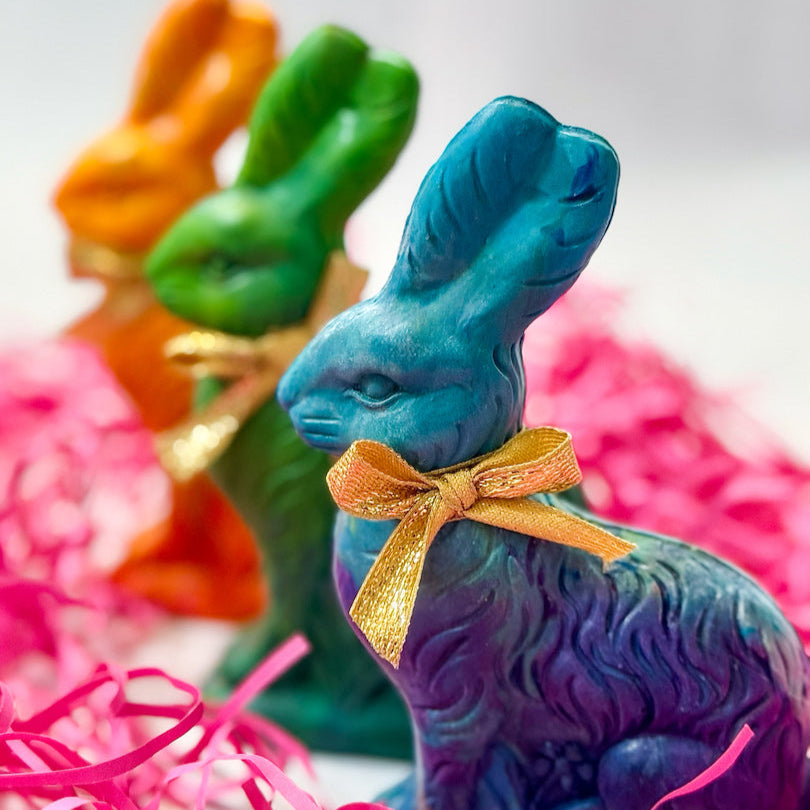 Three Easter Bunny crayons for kids in vibrant blue, green, and orange hues, each adorned with a gold ribbon. These handmade bunny-shaped crayons are perfect for Easter basket gifts, party favors, and spring art activities. The colorful, vintage-style rabbits are displayed on a festive background with pink Easter grass, making them a fun and unique Easter basket fillers.