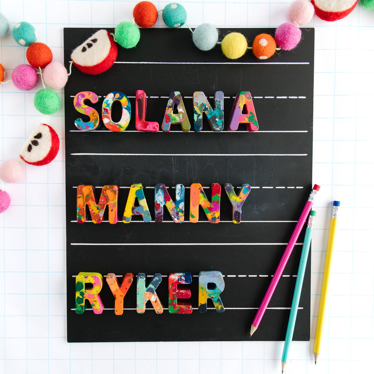 Back to school gift guide gift for kids. Three rainbow colored crayon names are displayed on a black chalkboard and a back to school themed apple garland borders the chalkboard as a photo prop. Rainbow Crayon names spell SOLANA, MANNY, and RYKER. 