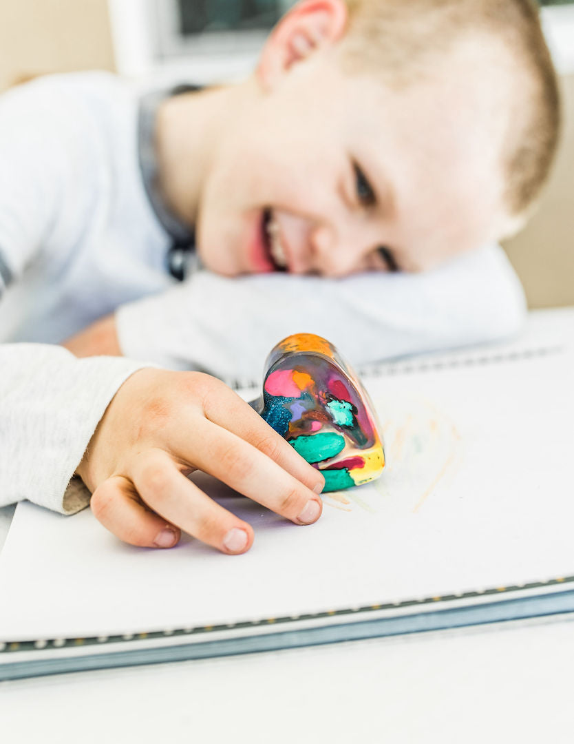 Boy holding heart shaped jumbo crayon from Art 2 the Extreme in his right hand on top of a sketch book. This heart crayon is multicolored and made from recycled crayons, crayon melting heart shape