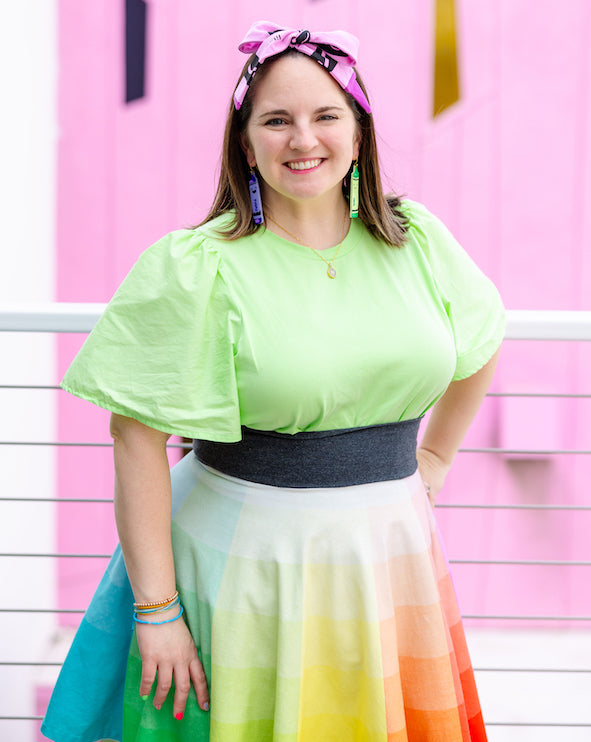 Women smiling in a lime green shirt and rainbow skirt that looks like a color wheel skirt. Rainbow crayon artist, Nicole Lewis, is standing outside on a pink wall and wears a purple crayon bow in her hair with day the crayons quit earrings.