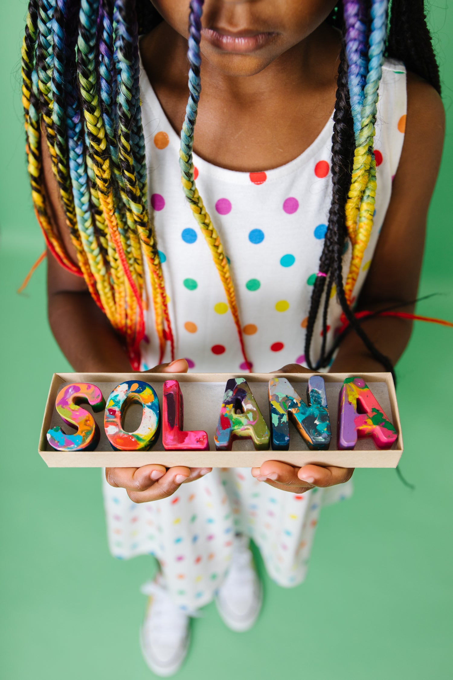Girl child with rainbow braids holds an open set of personalized name crayons that spell the name SOLANA in multicolored crayon letters. This crayon set from Art 2 the Extreme makes a unique birthday gift, holiday stocking stuffer, or goodie bag for children. 