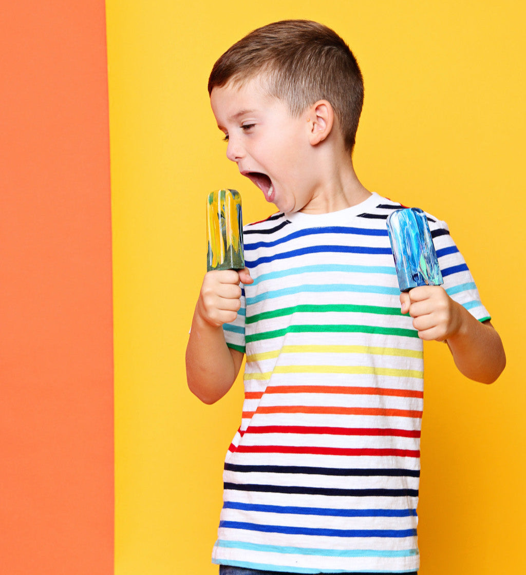 6-year-old boy with striped shirt holding two crayon popsicles, pretending he is going to eat one. Background is a solid yellow with a little orange to the viewers left. These crayons make great goodie bag favors for six year old! Shop at Art2theExtreme.com